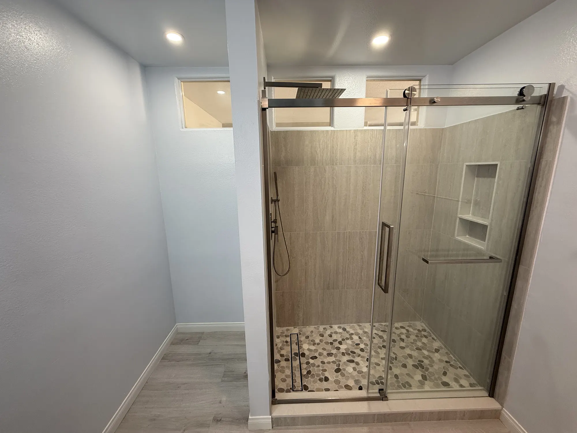 Finished walk-in shower with a frameless sliding glass door, pebble tile floor, tiled niche, rain head and handheld sprayer