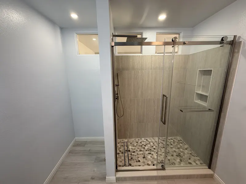 Finished walk-in shower with a frameless sliding glass door, pebble tile floor, tiled niche, rain head and handheld sprayer