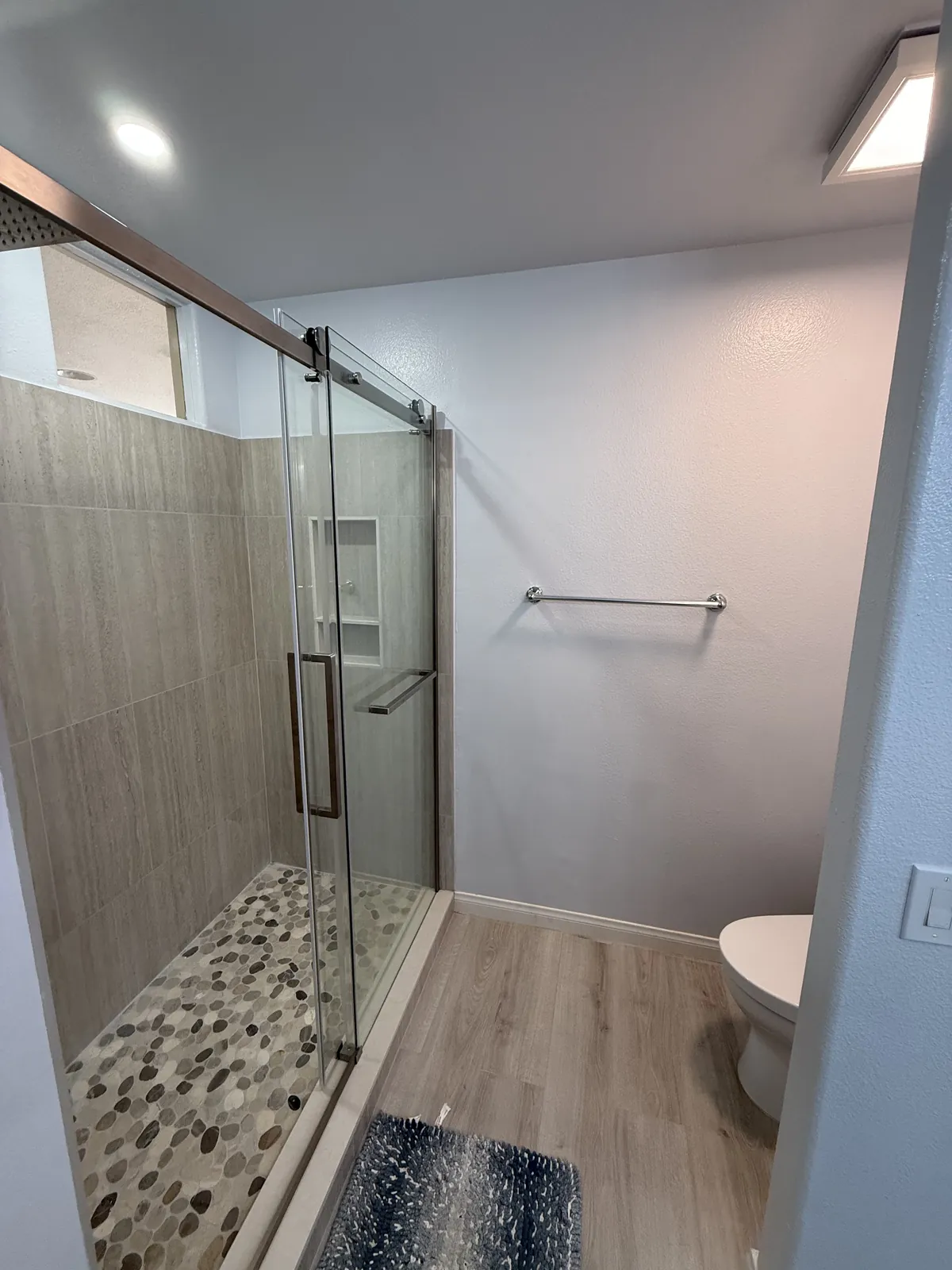 Angled view of the finished shower with pebble tile floor, sliding glass door, new plank flooring and a towel bar