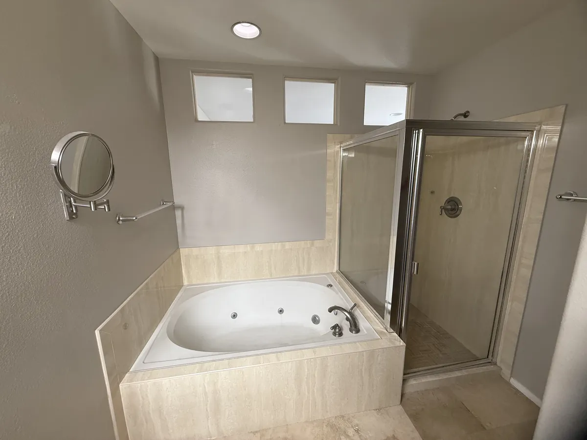 Bathroom before the remodel with a beige tile jetted tub under three small windows and a corner shower with a framed door