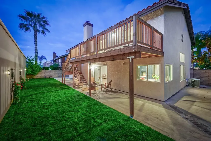 Finished backyard at dusk with a new second-story wood deck and stairs, covered patio, fresh lawn and lit kitchen window