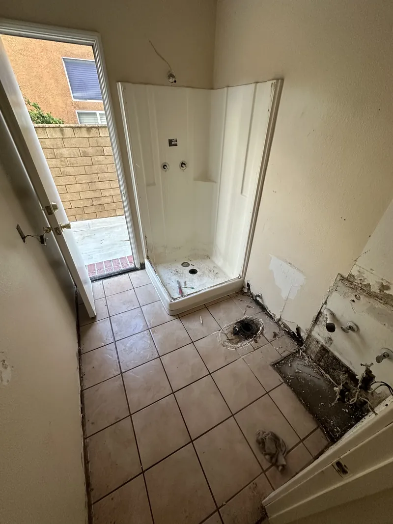 Old bathroom with a molded shower stall, beige floor tile and exposed plumbing where the toilet and vanity were removed