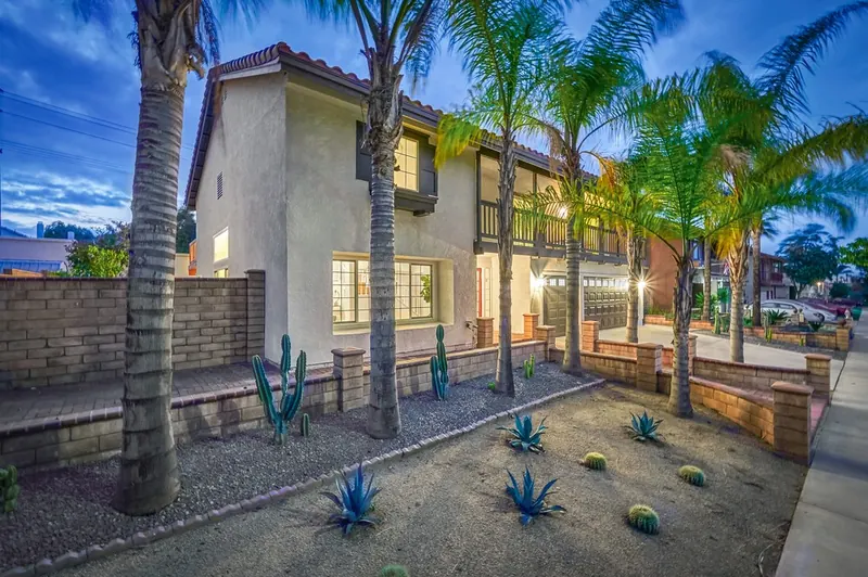 Repainted two-story home at dusk from the front corner with a wood balcony, palms and gravel planters with agave