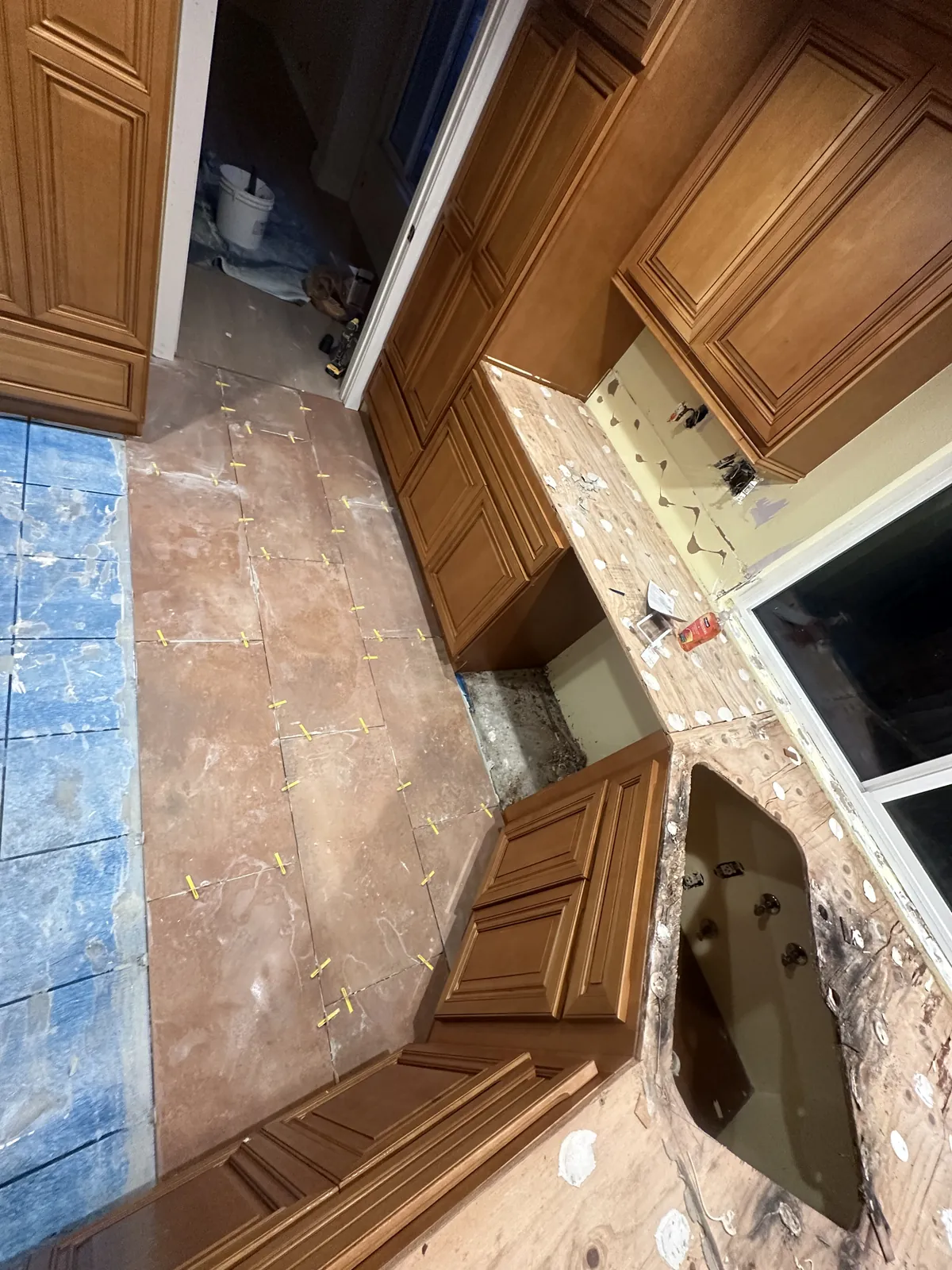New terracotta-tone floor tile with spacers going down over the old blue kitchen tile, between wood cabinets