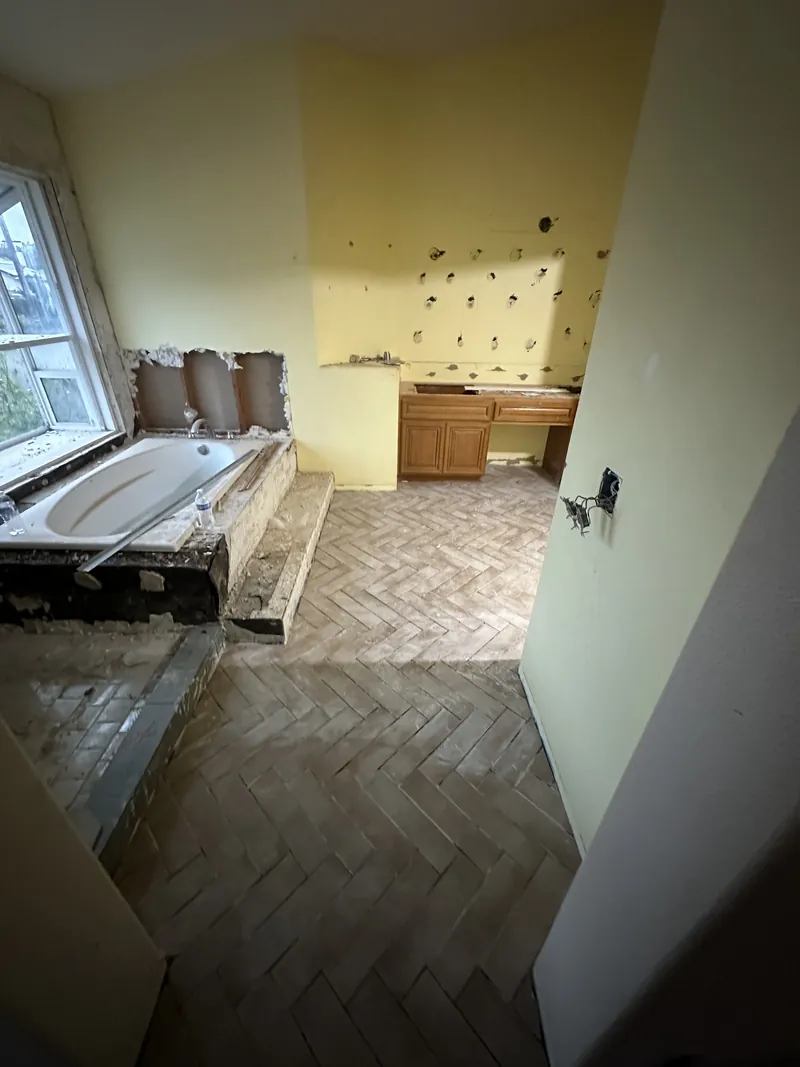 Primary bathroom mid remodel with the old tub deck stripped, new herringbone floor laid and the original vanity beyond