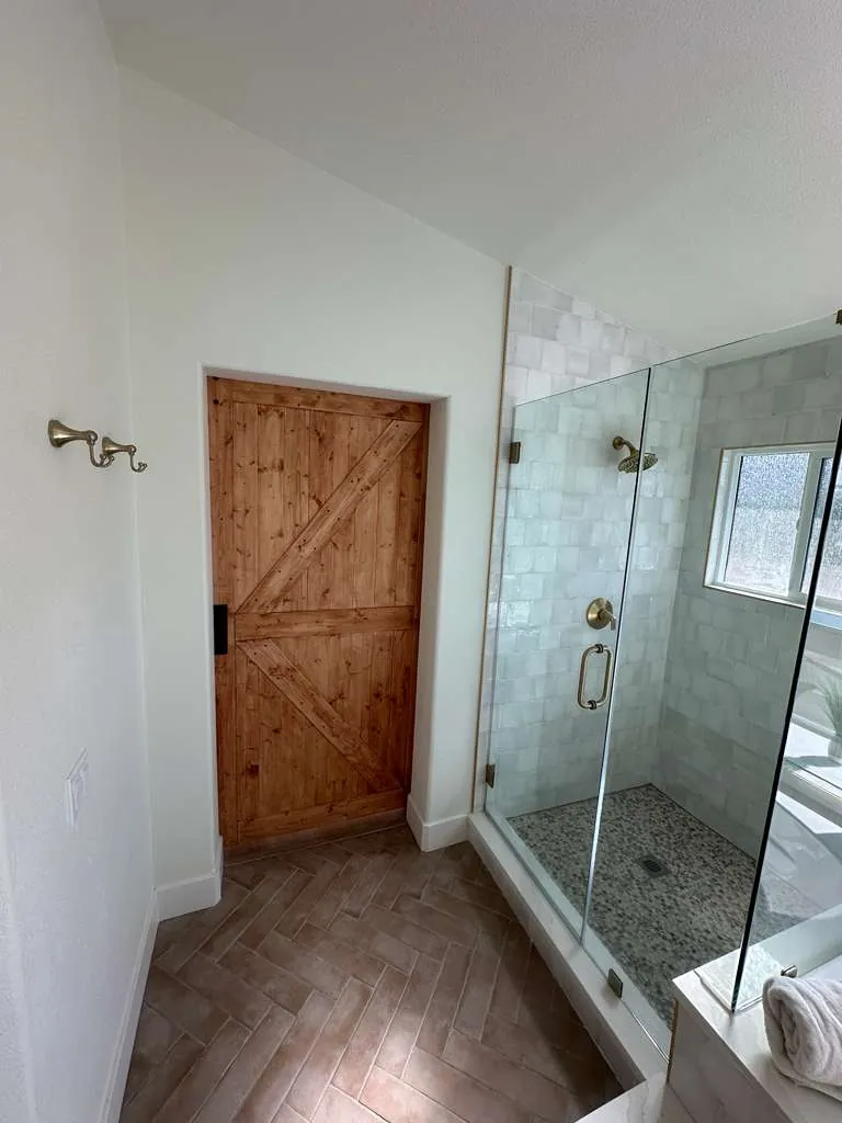 Frameless glass shower with glossy white square tile, pebble floor and brass-tone fixtures beside a knotty wood barn door