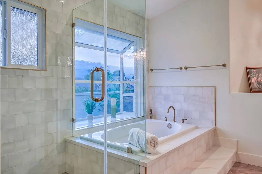 Tiled soaking tub under a garden window with brass-tone fixtures, seen past the frameless glass shower enclosure