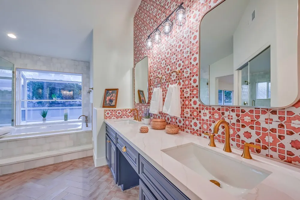 Navy double vanity with a white veined top and brass-tone faucets against a red patterned tile wall and cage lights