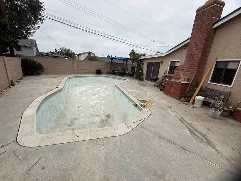 Wide view of the drained backyard pool with old stained plaster, concrete deck and house before the work