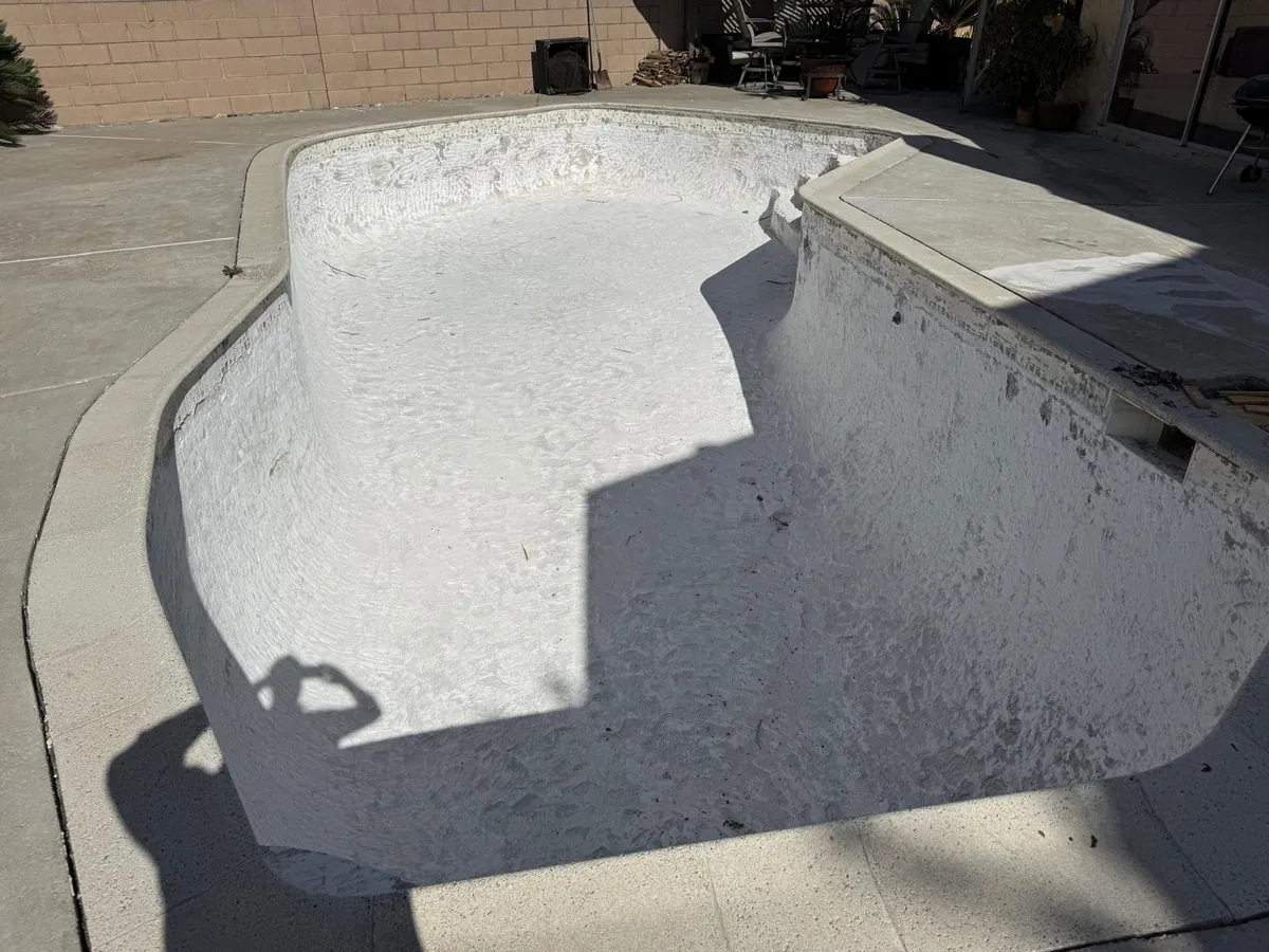 Empty pool basin coated in fresh white plaster before refilling, with the concrete deck and block wall around it