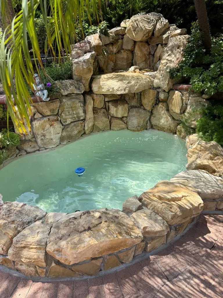Refilled backyard fountain with clear turquoise water under a stacked rock waterfall, ringed by rock and a brick patio