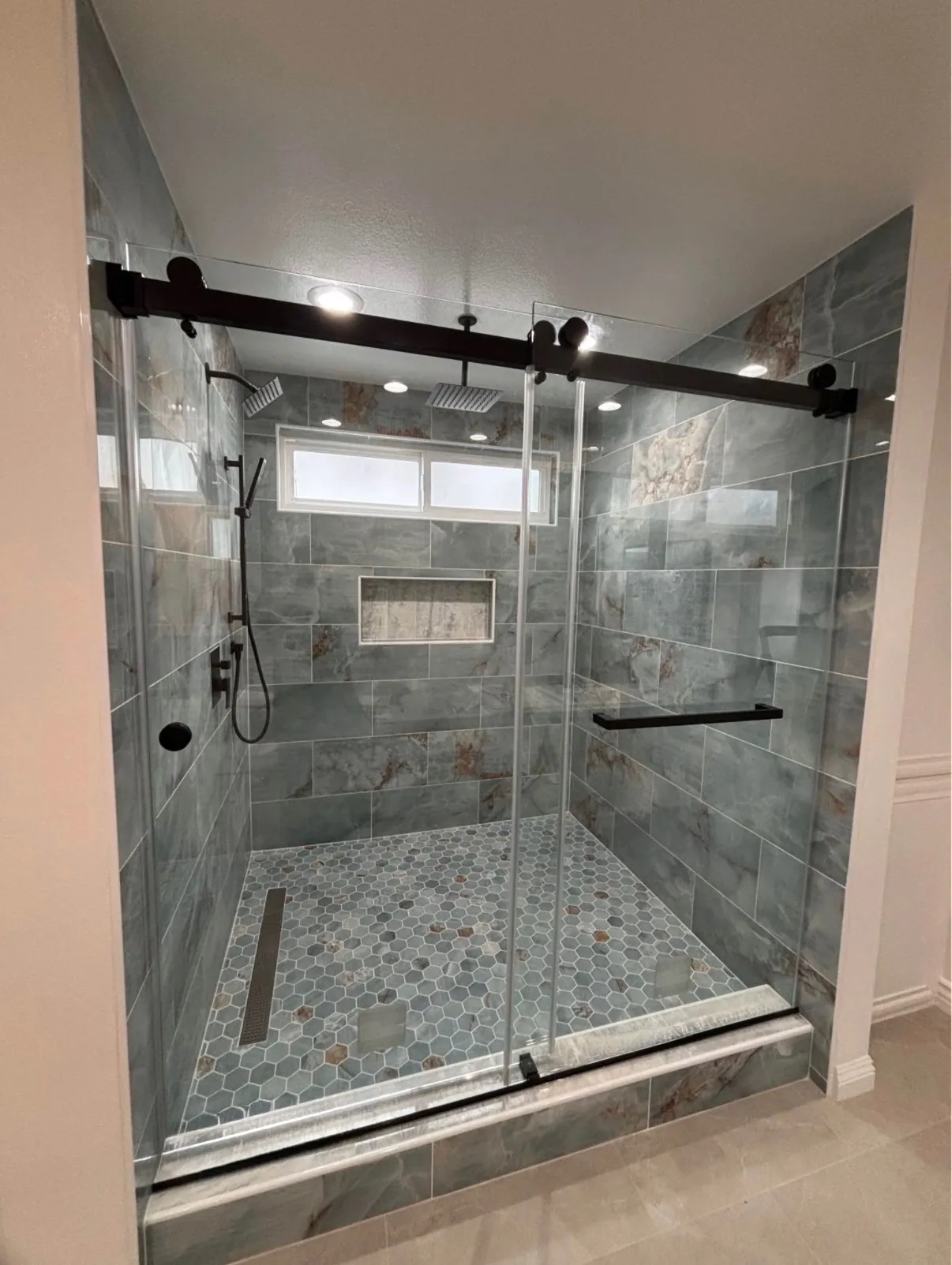 Walk-in shower with sliding glass on black track hardware, green-gray marble-look tile and a mosaic floor
