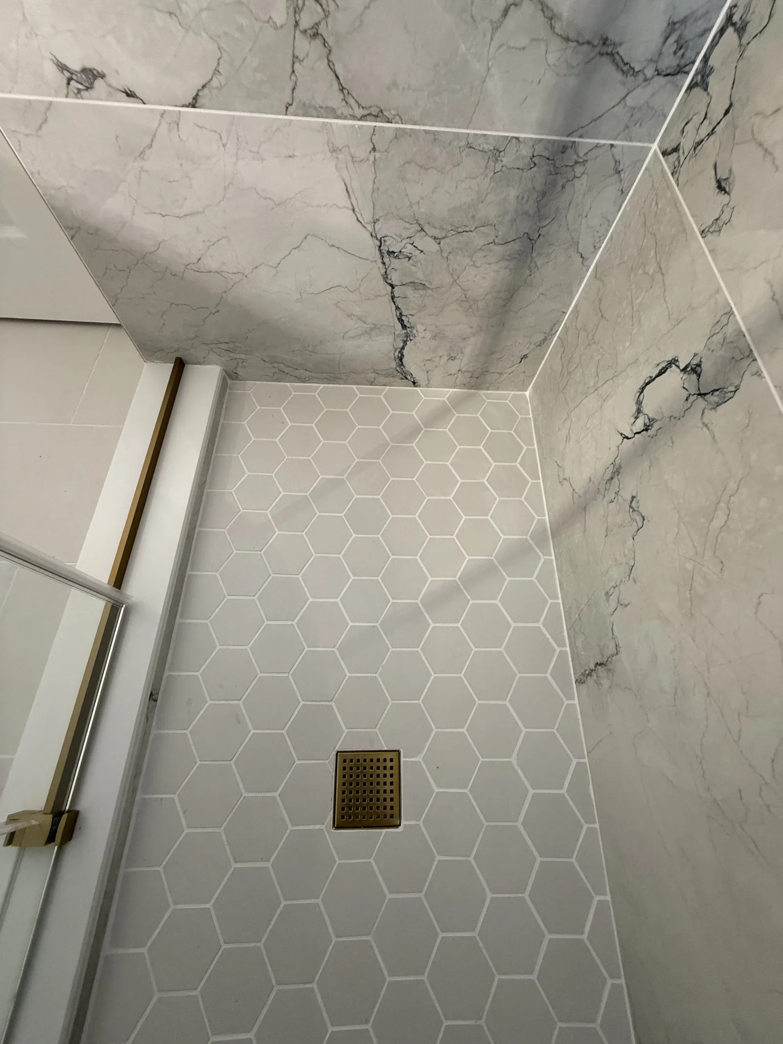 Shower lined floor to ceiling in marble-look slab with a white hexagon tile floor and a square gold rain head