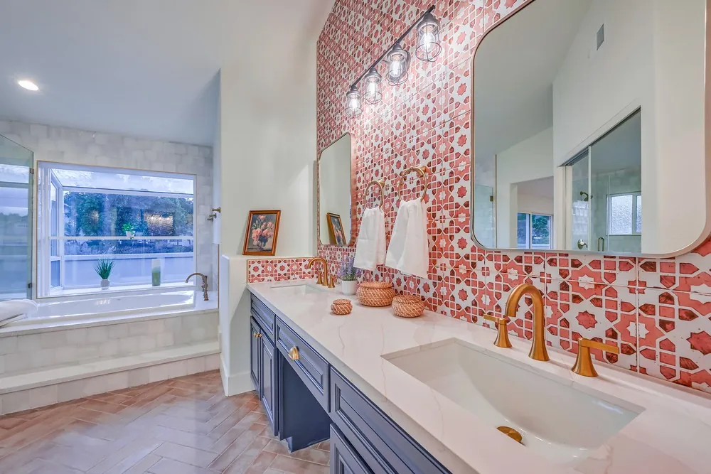 Double vanity with navy cabinets, a white counter, gold faucets and a patterned red tile wall between two mirrors