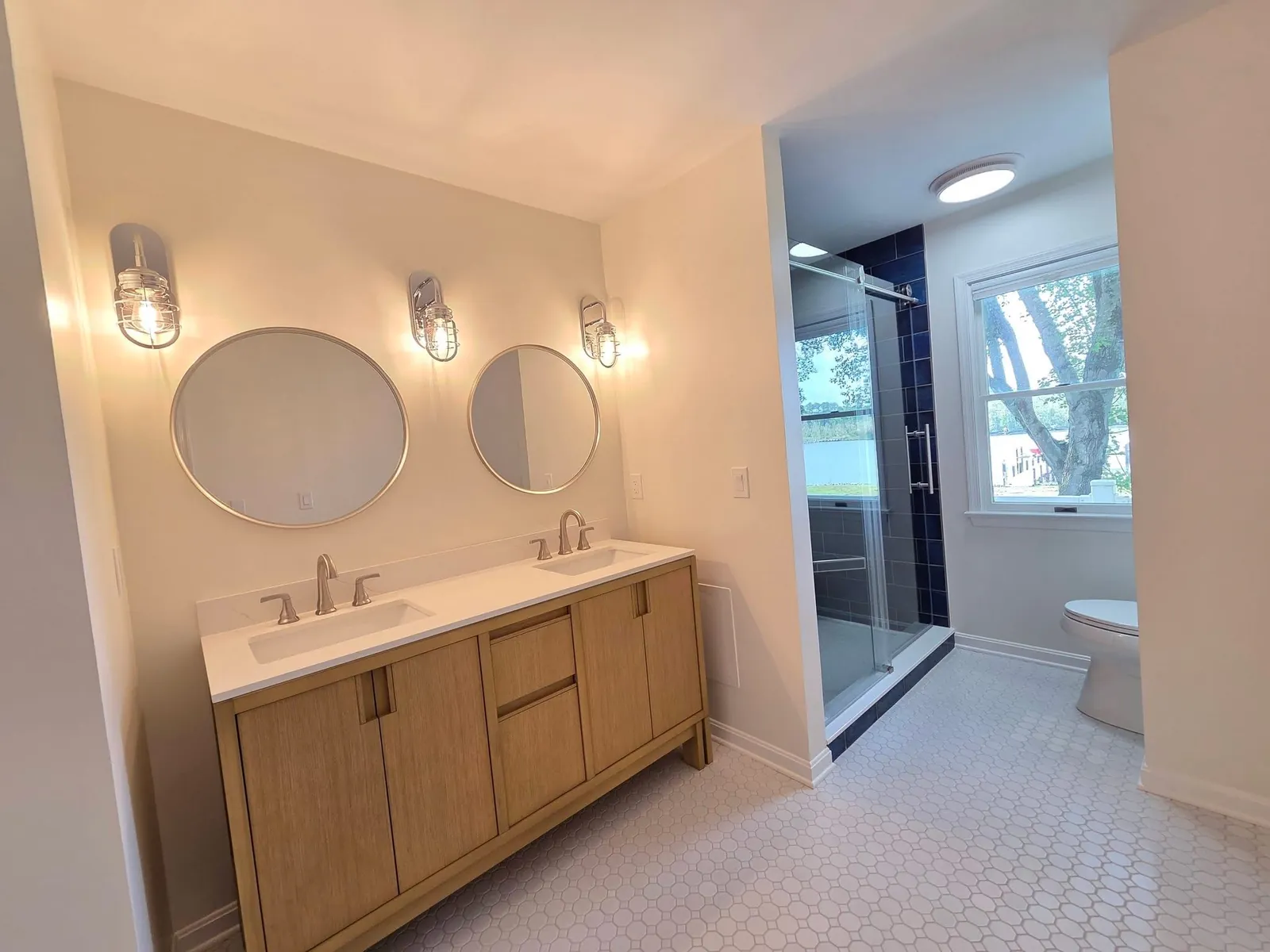 Double vanity in light oak with two round mirrors, wall sconces and a hexagon tile floor, with the shower beyond