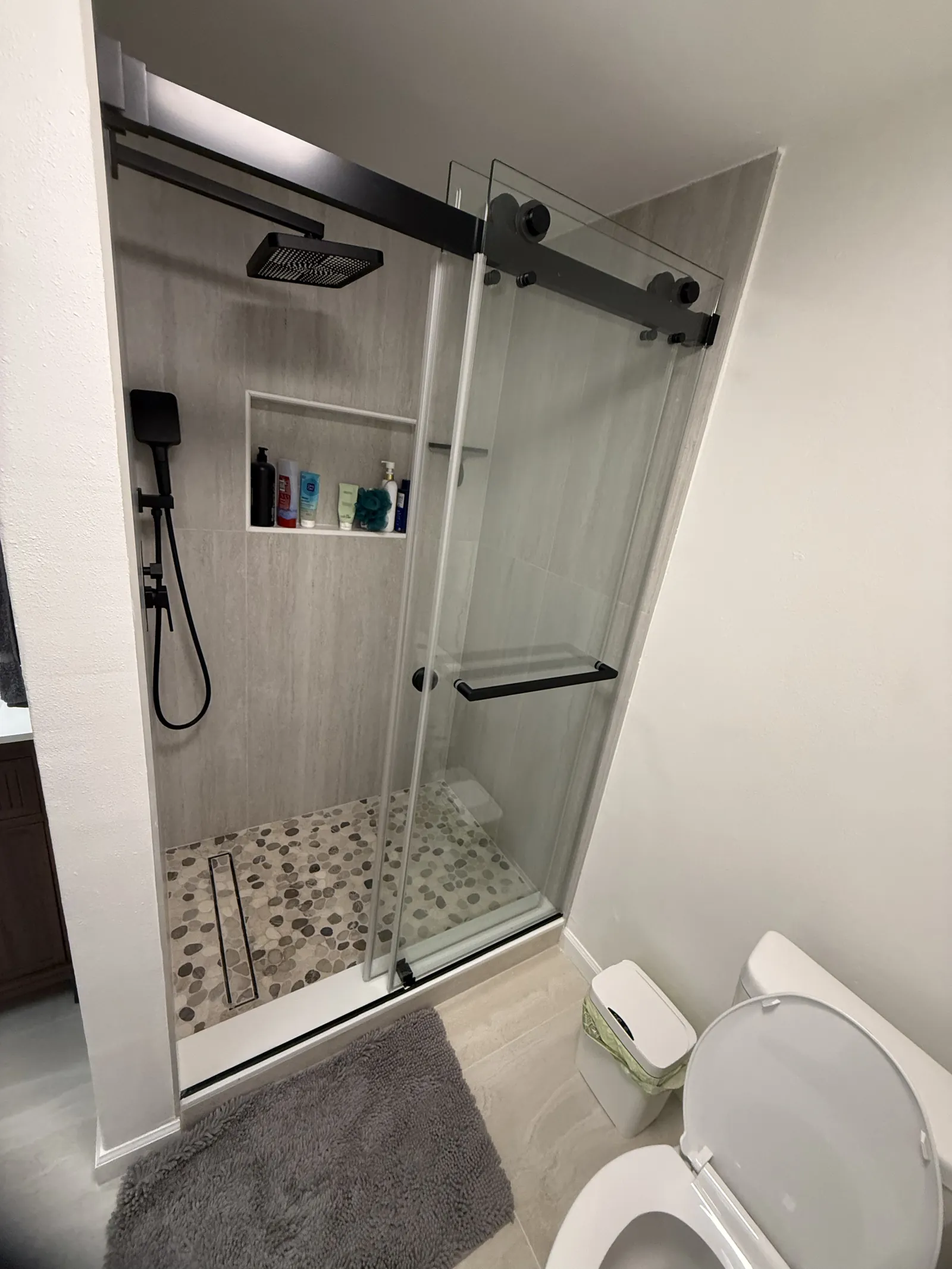 Shower behind sliding glass on black track hardware with gray tile, a pebble floor and black fixtures
