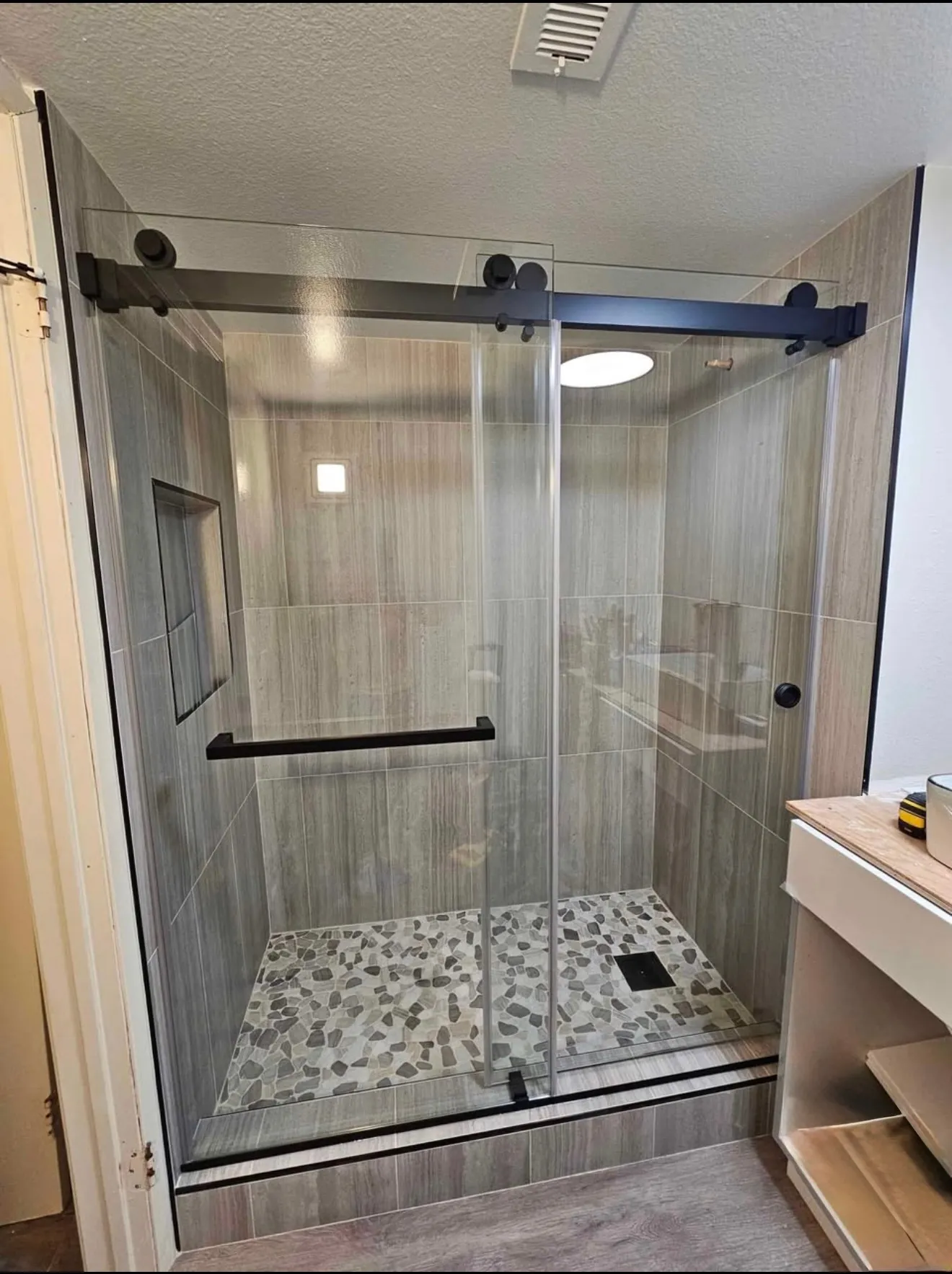 Shower behind sliding glass on black track hardware with wood-look tile, a pebble floor and a corner shelf