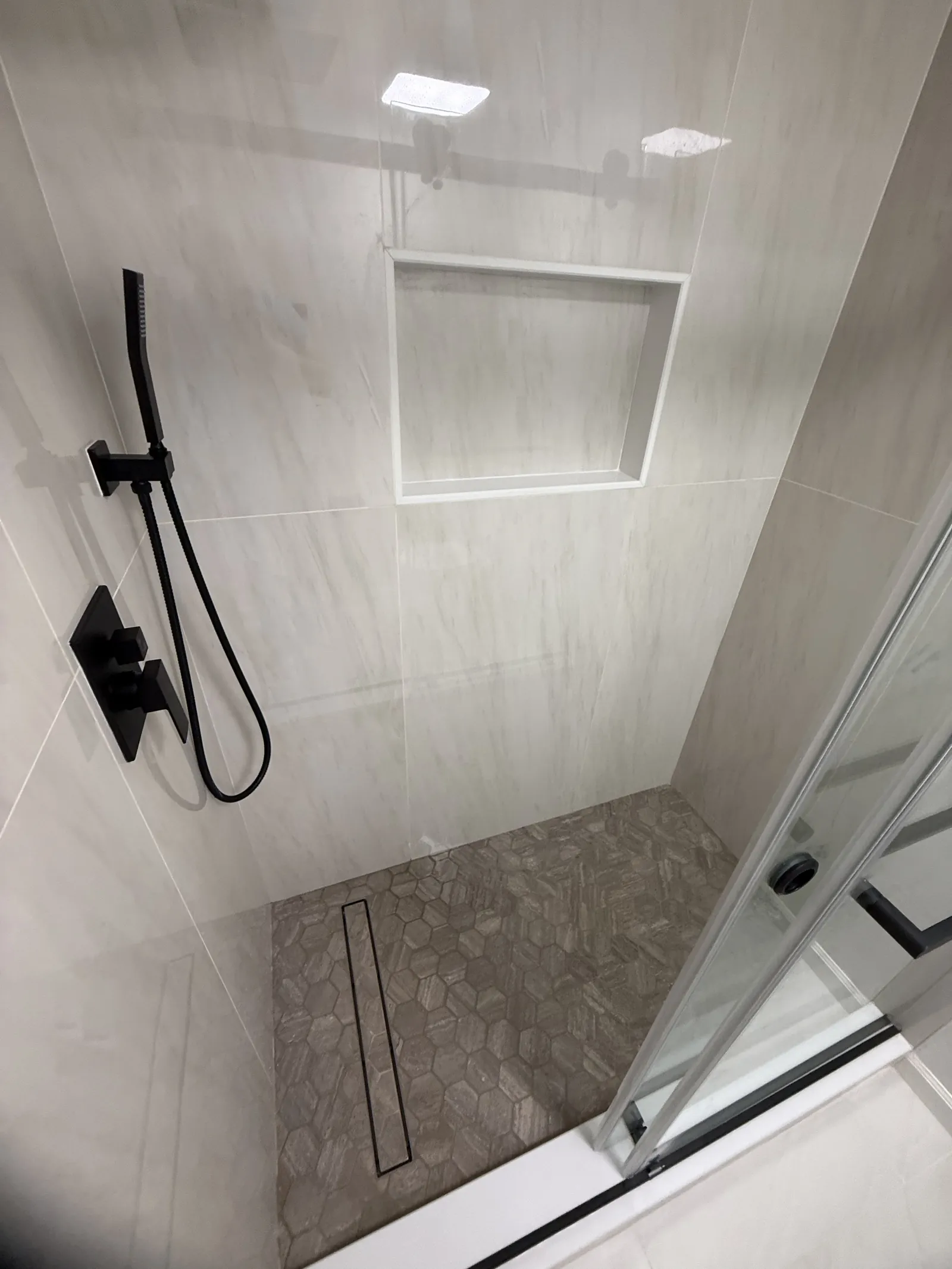 Shower in light stone-look tile with black fixtures, a pebble floor and a linear drain