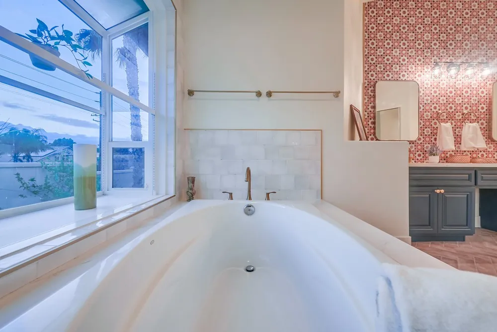 Soaking tub under a window with marble tile surround and a patterned red tile wall behind the vanity