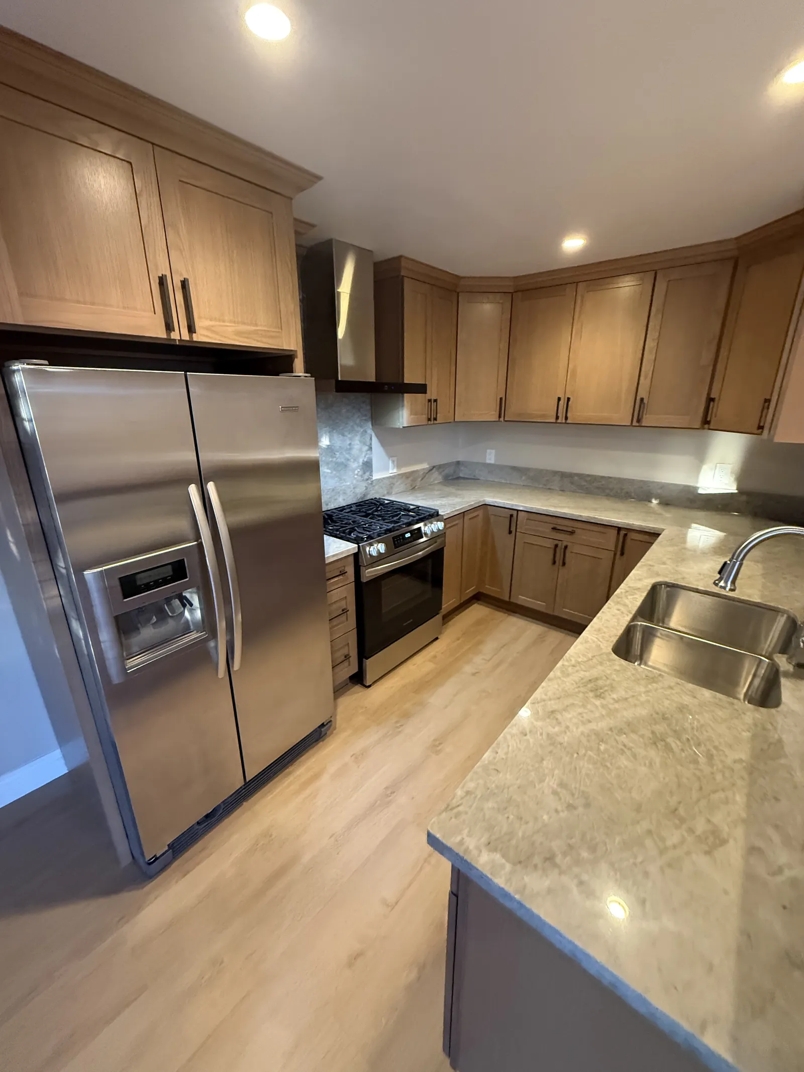 Corner of an oak kitchen with a stainless fridge and range, a stone counter and an undermount sink